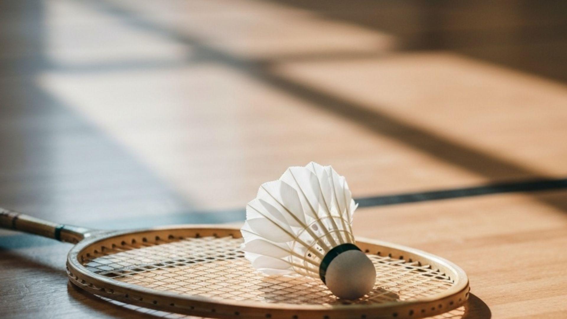 En träbadmintonracket och en fjäderboll vilar på ett blankpolerat sportgolv i mjukt sidoljus